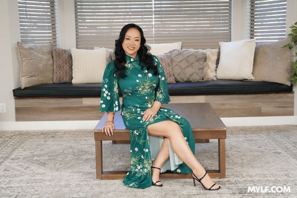 Asian woman in a green floral dress sits on a wooden table, smiling. She is indoors with a bench and several decorative pillows behind her, and there are large windows with blinds in the background.