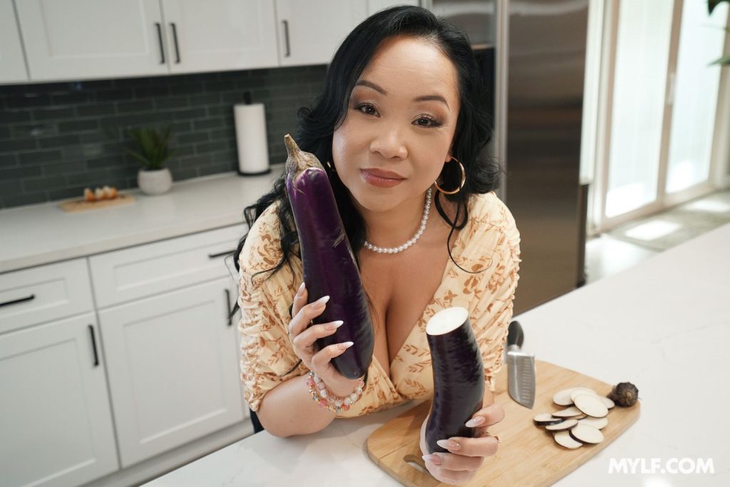 Asian woman in a kitchen holds a whole eggplant and a cut eggplant. Sliced pieces are on a cutting board with a knife. She looks at the camera and smiles slightly. White cabinets and a green tile backsplash are in the background.