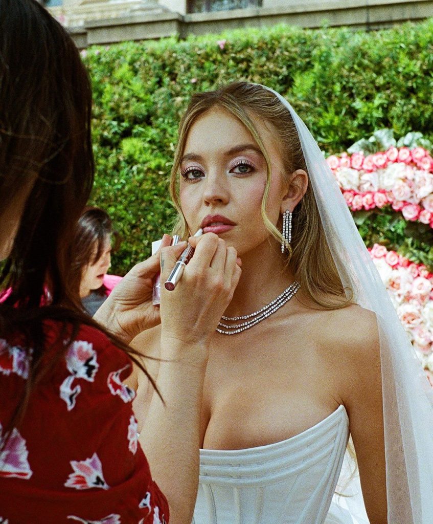 A bride in a strapless white dress and veil has her makeup touched up outdoors. She’s wearing diamond jewelry and standing near a hedge and pink flower arrangement. Another person applies lipstick to her lips.