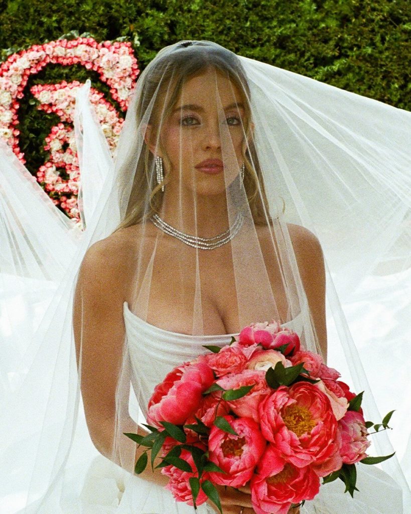 A bride in a white dress and veil holds a bouquet of vibrant pink flowers. She has long hair, wears silver jewelry, and stands outdoors in front of green bushes and a pink floral heart decoration.