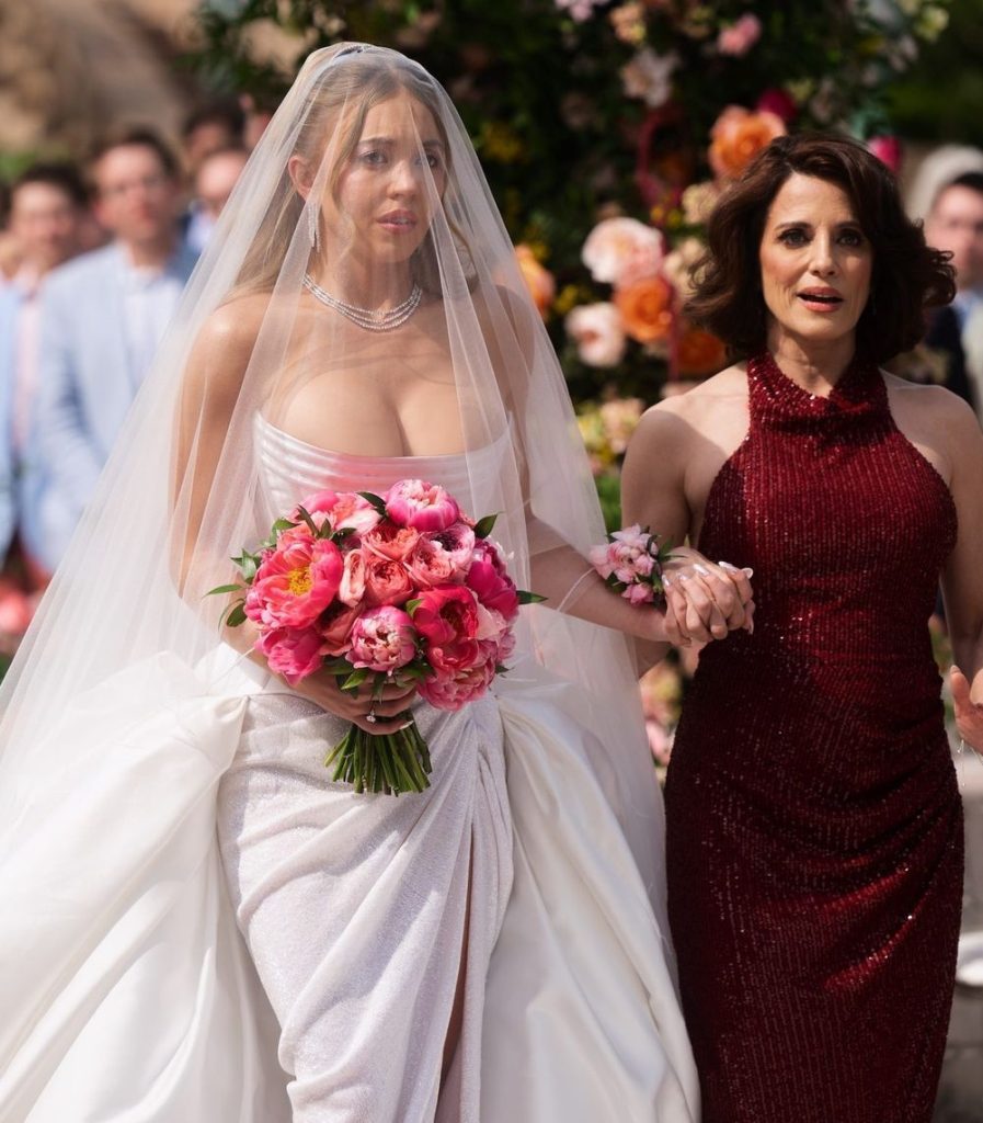 A bride in a white dress and veil, holding a bouquet of pink flowers, walks down the aisle holding hands with a woman in a sleeveless red gown; guests watch in the background.
