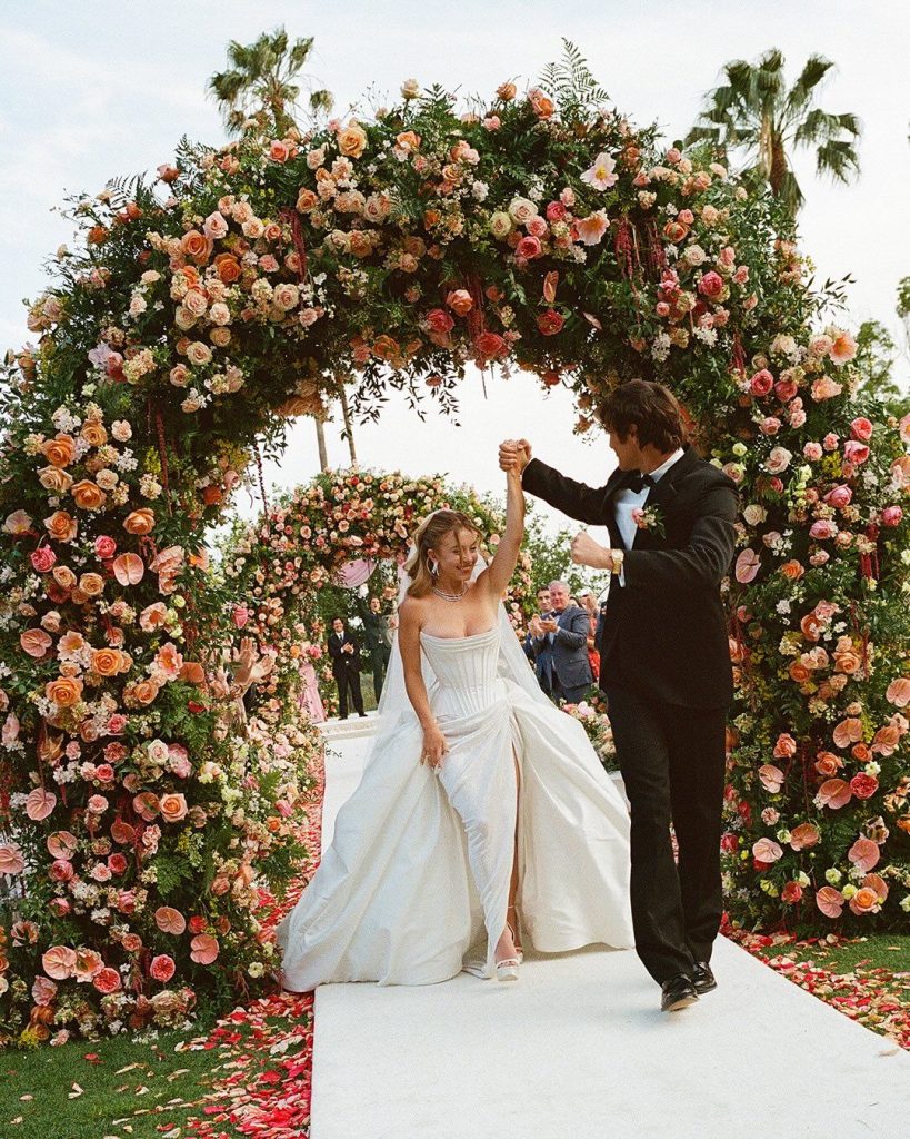A bride in a white dress and a groom in a black suit walk under a large floral archway, surrounded by lush arrangements of colorful flowers and palm trees, smiling and holding hands during an outdoor wedding.