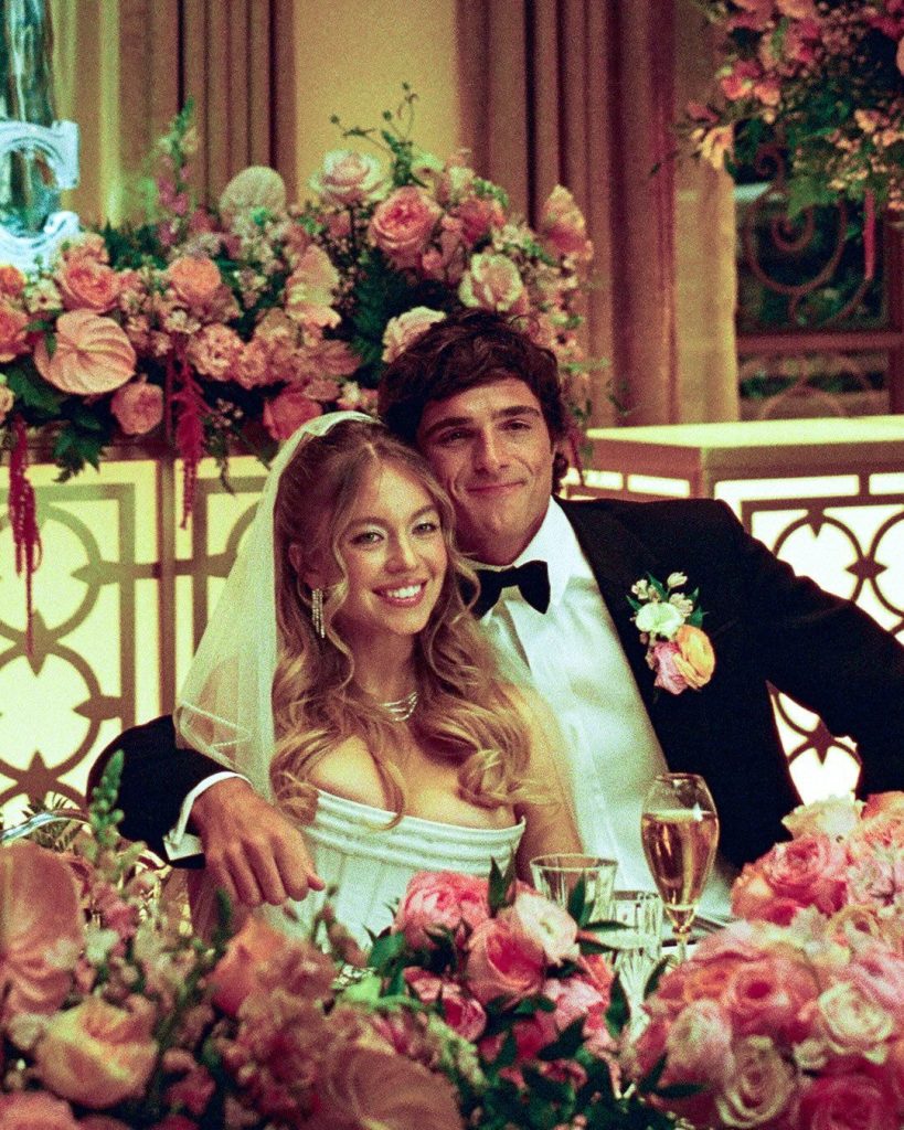 A bride in a white dress and veil sits beside a groom in a black tuxedo, both smiling, surrounded by pink and peach flowers at a decorated wedding reception table.