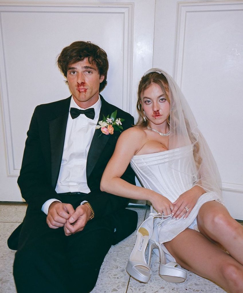 A bride and groom in formal wedding attire sit on the floor against a wall, both with blood on their faces and clothes. The groom wears a tuxedo; the bride wears a white dress, veil, and holds high-heeled shoes.