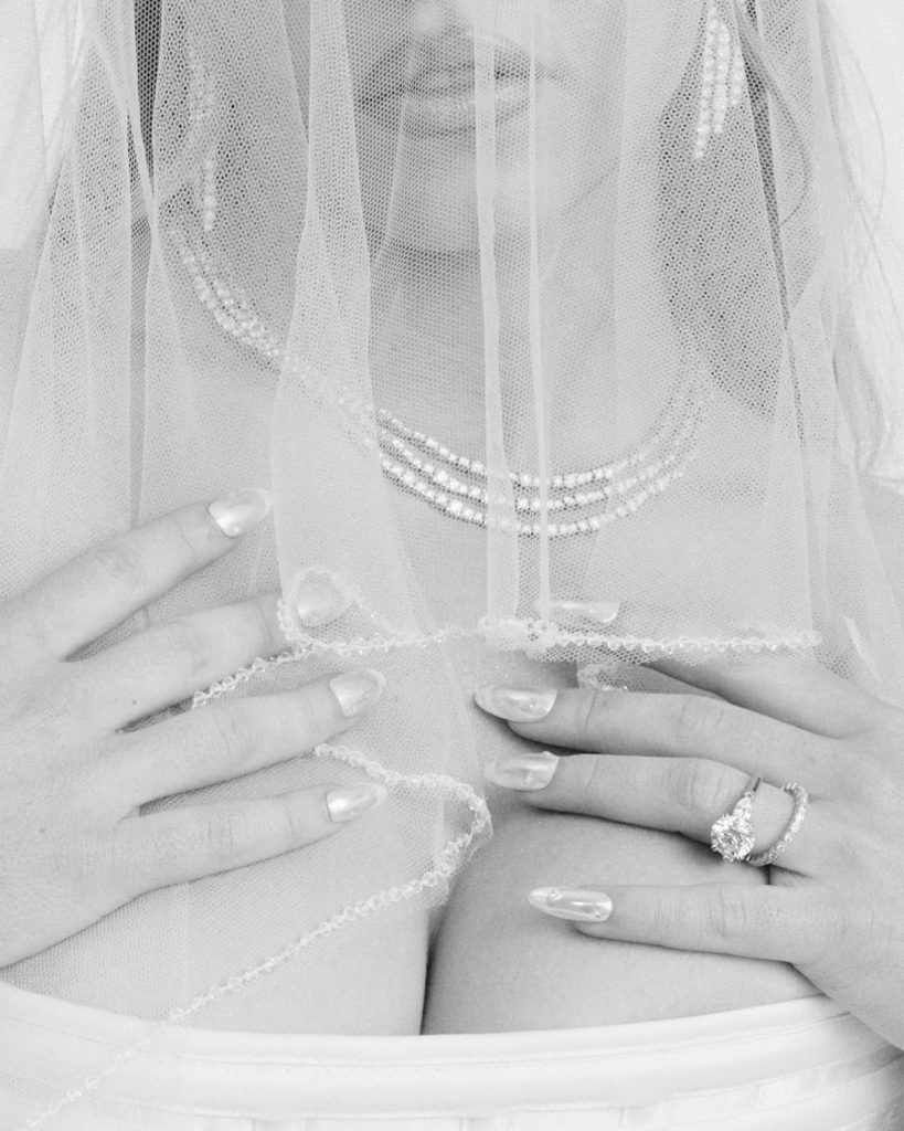 A close-up black-and-white photo of a bride with a veil covering her face, wearing a pearl necklace and earrings, showing manicured nails and a wedding ring, with her hands resting on her chest.