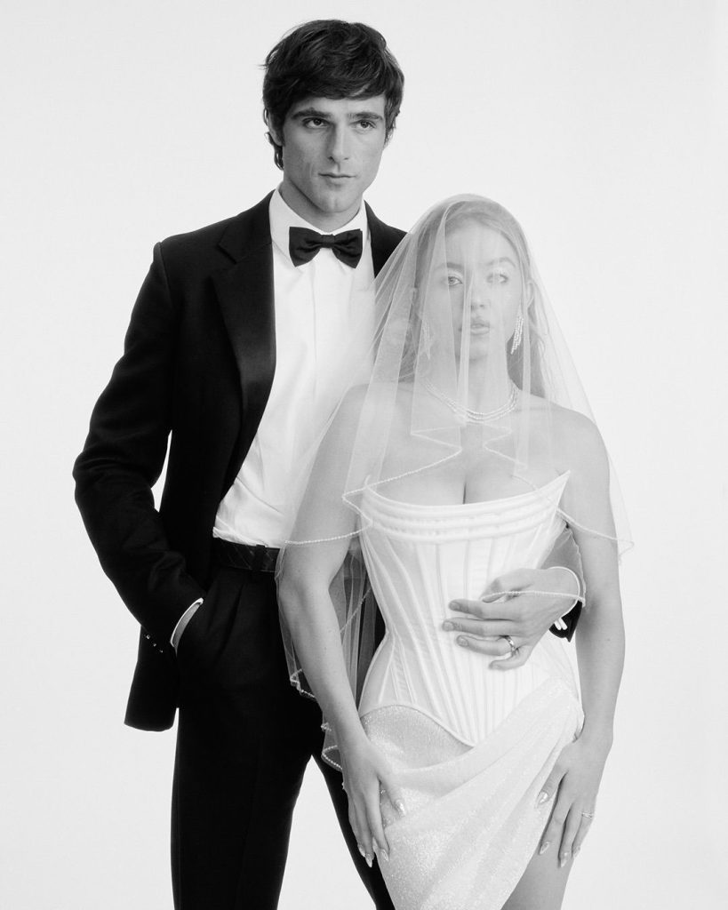A man in a tuxedo stands beside a woman wearing a strapless corset dress and a veil. The man has one arm around her waist. The image is black and white, with a minimal studio background.