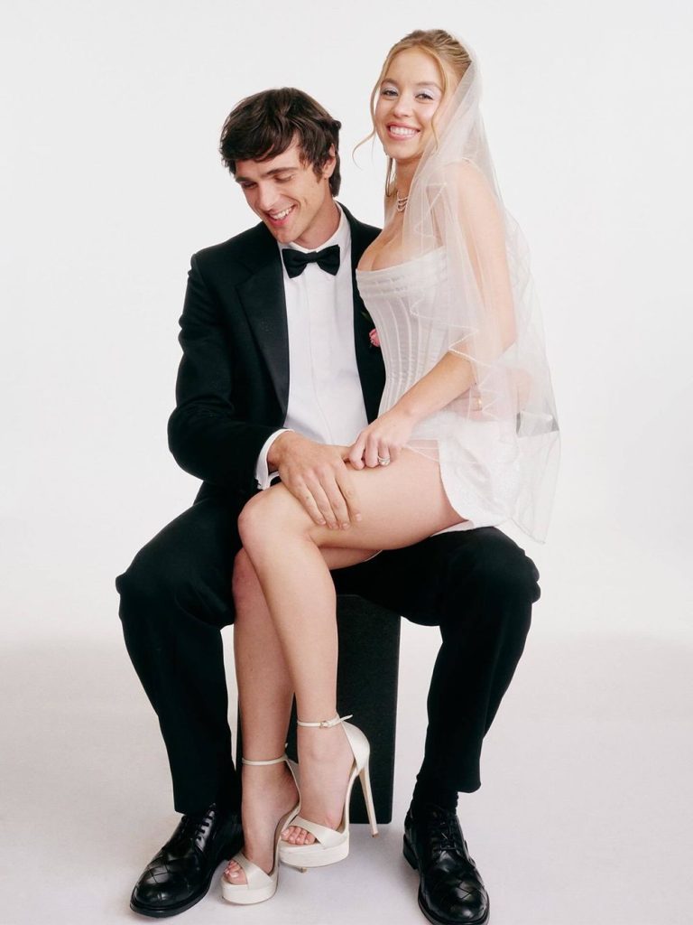 A young couple poses for a wedding photo. The man wears a black tuxedo and bow tie, sitting on a stool, while the woman in a short white dress, veil, and high heels sits on his lap, smiling. The background is plain white.