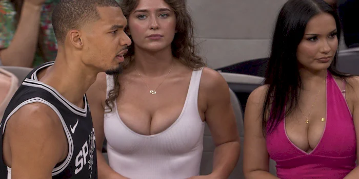 A basketball player in a black Spurs jersey stands on the court. Behind him, two women with long hair sit in the audience, one in a white top and the other in a pink top, both looking ahead.