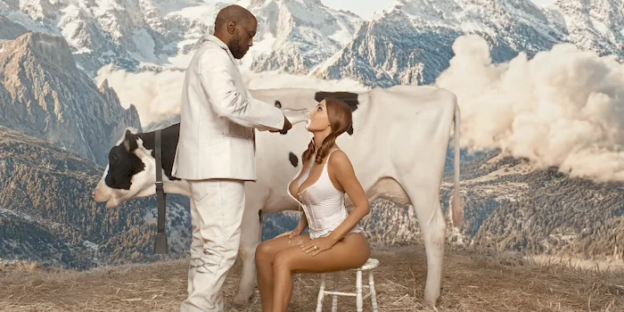 A man in a white suit stands in front of a seated woman in a revealing outfit beside a cow, with a backdrop of snowy mountains and clouds. The scene has a surreal, staged appearance.