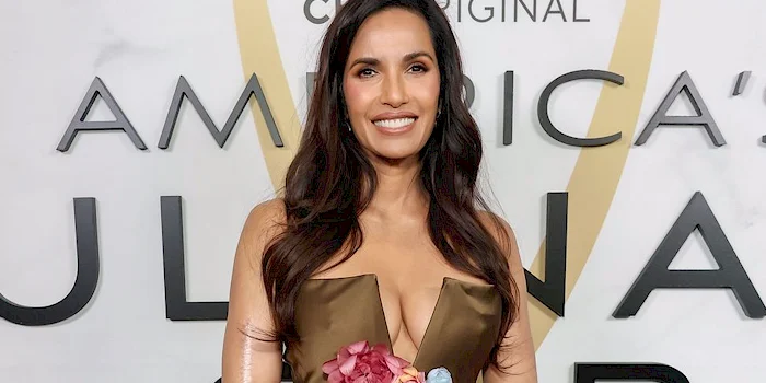 Close up of a smiling Padma Lakshmi wearing a strapless dress in front of a white background with big text on it.