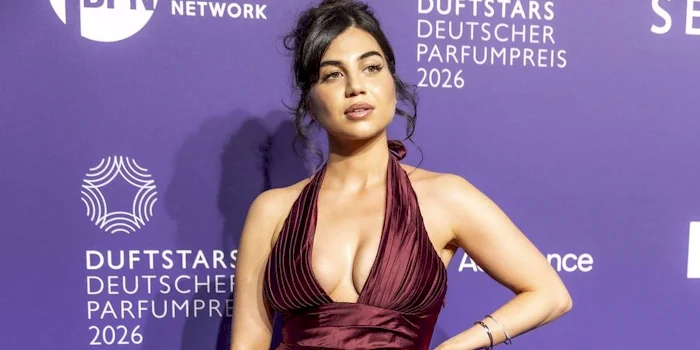 A woman in a deep burgundy, halter-neck gown poses in front of a backdrop with various sponsor logos and event details.
