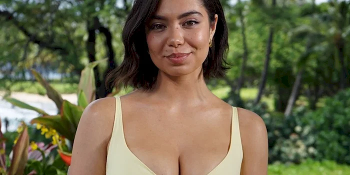 Close up of brunette woman wearing a yellow dress and looking into the camera with a slight smile. Greenery is in the background.