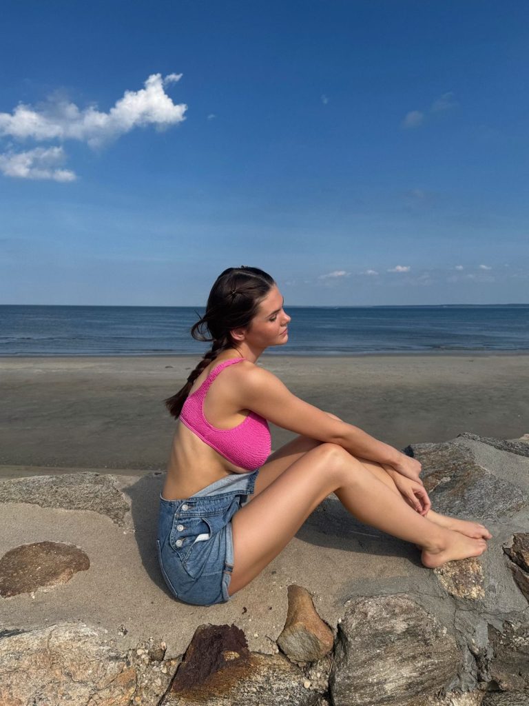 A woman in a pink top and denim overalls sits barefoot on rocks by a sandy beach, facing the ocean under a blue sky with a few clouds.