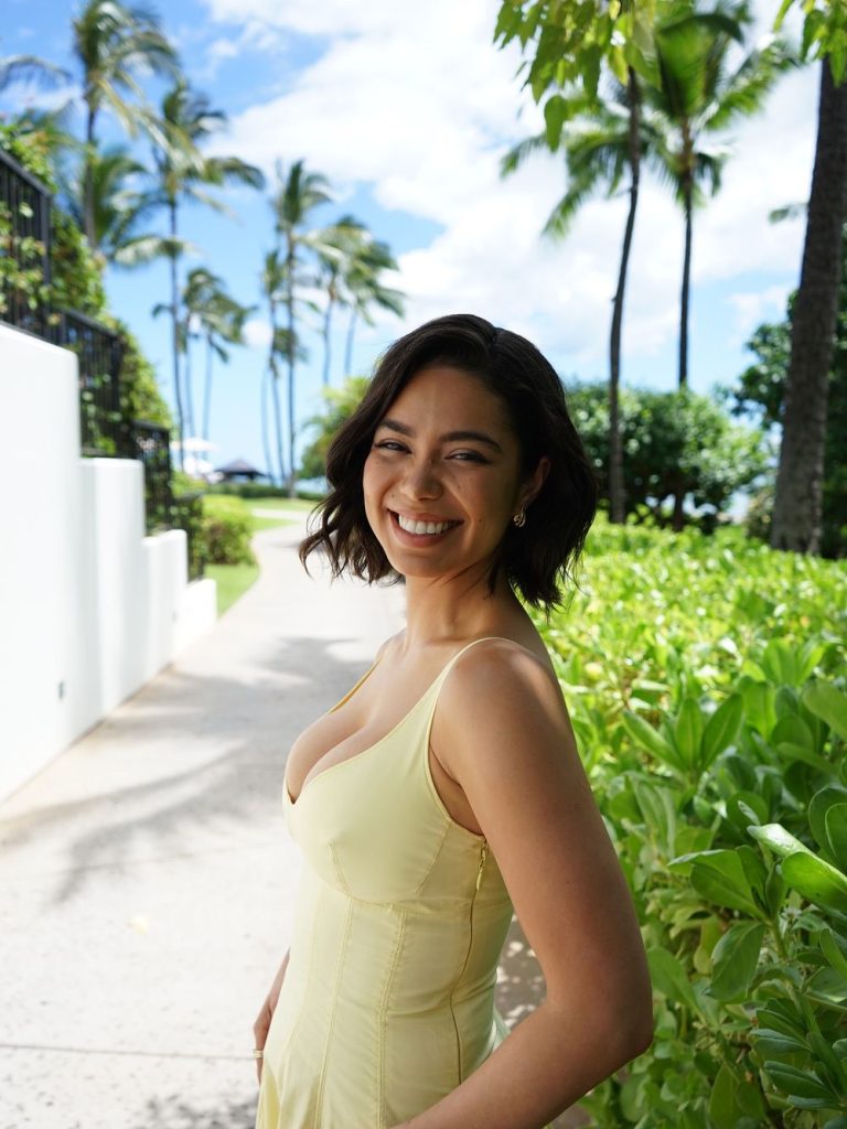 A woman with short dark hair in a sleeveless yellow dress smiles while standing on a sunlit path surrounded by green bushes, palm trees, and a blue sky with some clouds.
