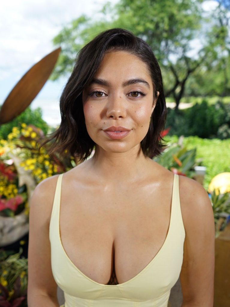A woman with short brown hair wearing a yellow sleeveless top stands outdoors in front of colorful flowers and green trees, looking directly at the camera with a neutral expression.