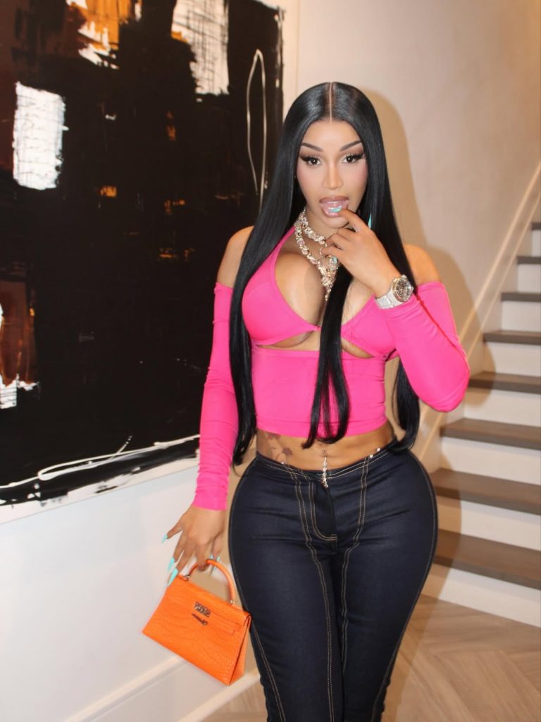 A woman with long straight black hair wears a bright pink cut-out top and dark high-waisted jeans, accessorized with layered necklaces, a watch, and an orange mini handbag, standing indoors near a staircase and abstract art.