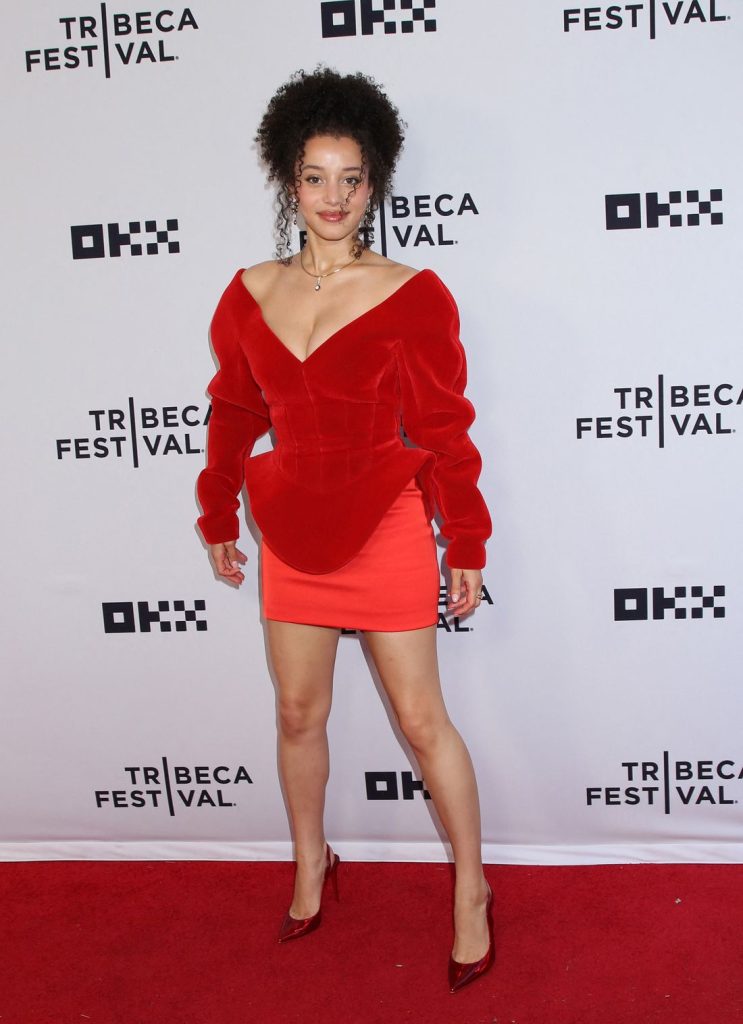 A woman with curly hair stands on a red carpet at the Tribeca Festival, wearing a red off-the-shoulder dress with structured shoulders and matching red heels, posing in front of a white backdrop with festival logos.