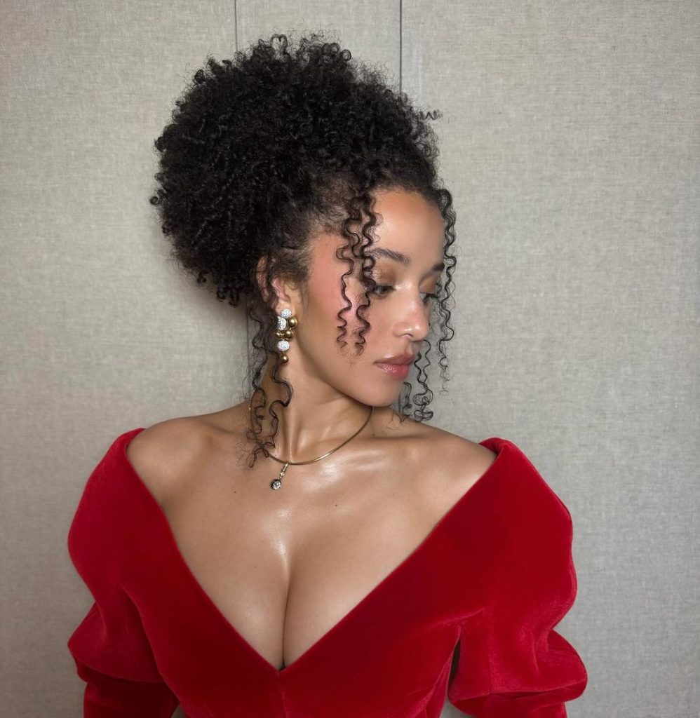 A woman with curly hair styled in an updo wears a red, off-the-shoulder dress with a deep neckline. She has pearl earrings and a necklace, and she is looking down to her left against a plain background.