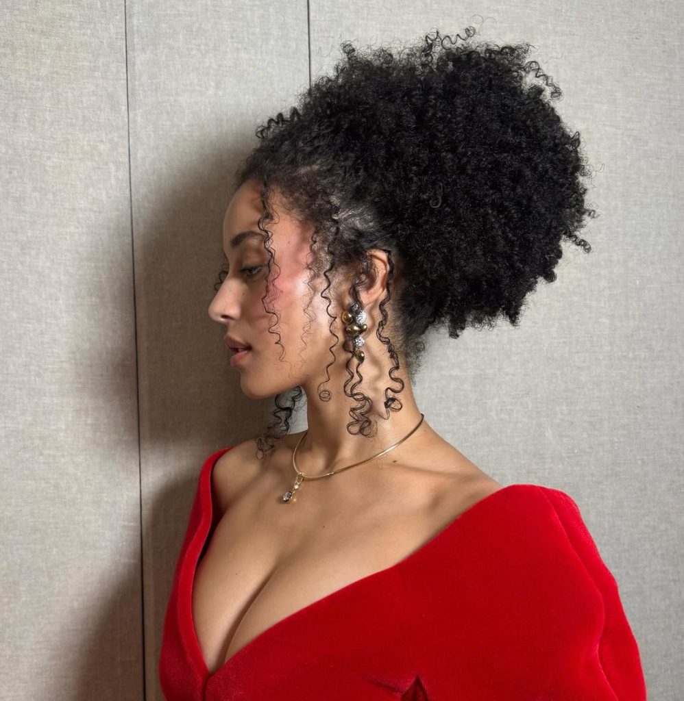 A woman with curly hair in a puff hairstyle wears a red off-the-shoulder dress, gold necklace, and pearl earrings, standing against a light-colored textured wall, looking to the left.