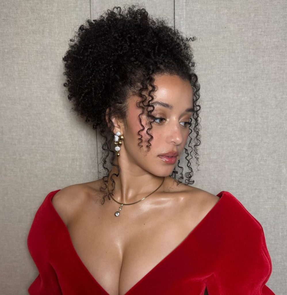 A woman with curly dark hair styled up, wearing a red off-the-shoulder dress, pearl earrings, and a delicate necklace, poses against a neutral background, looking to the side.