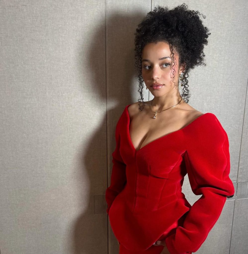 A woman with curly hair wearing a bold red, off-the-shoulder dress stands against a light-colored wall, looking to the side with a calm expression.