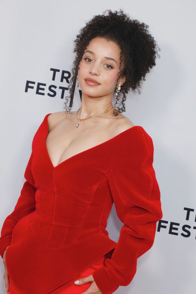 A woman with curly hair poses in a bold red off-the-shoulder dress against a white backdrop with partial TRIBECA FESTIVAL text visible. She wears pearl earrings and looks confidently at the camera.