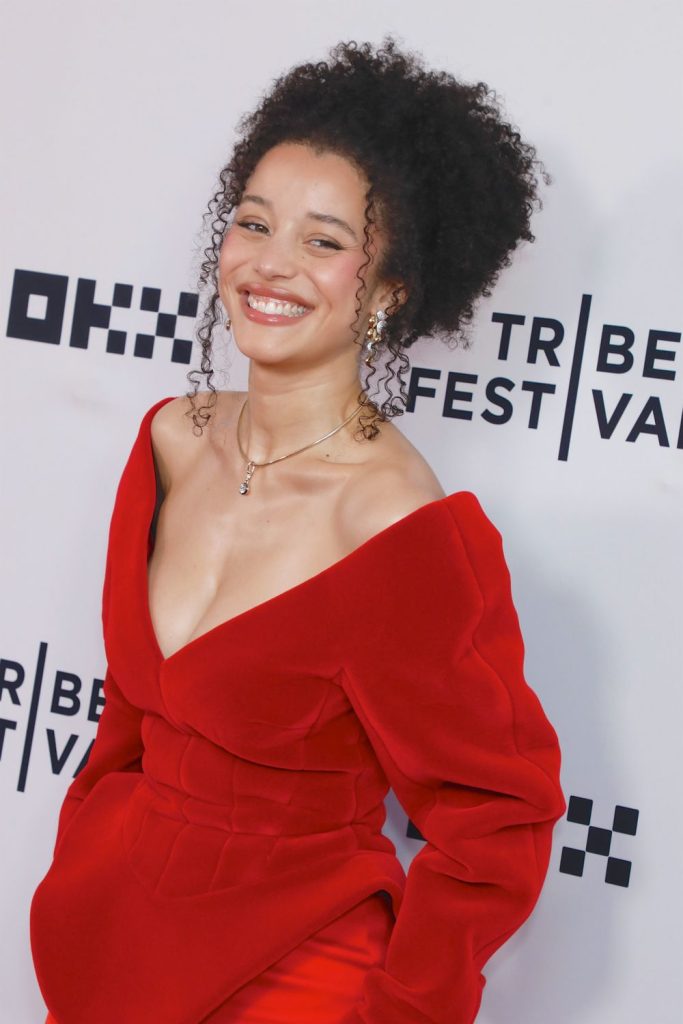 A woman with curly hair, wearing a bright red off-shoulder dress and a necklace, smiles while posing in front of a step-and-repeat backdrop with the Tribeca Festival logo.