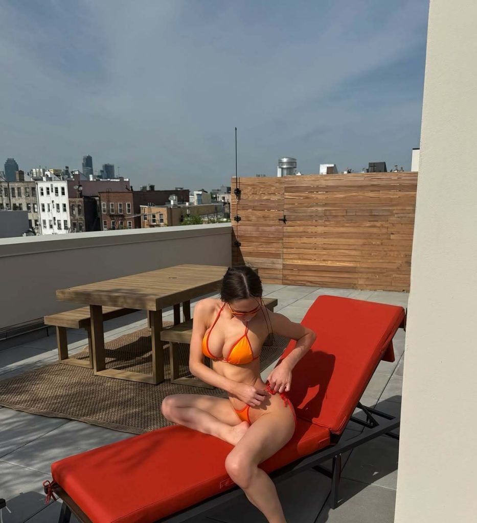 A woman in an orange bikini sits on a red lounge chair on a rooftop patio with wooden furniture and city buildings visible in the background.