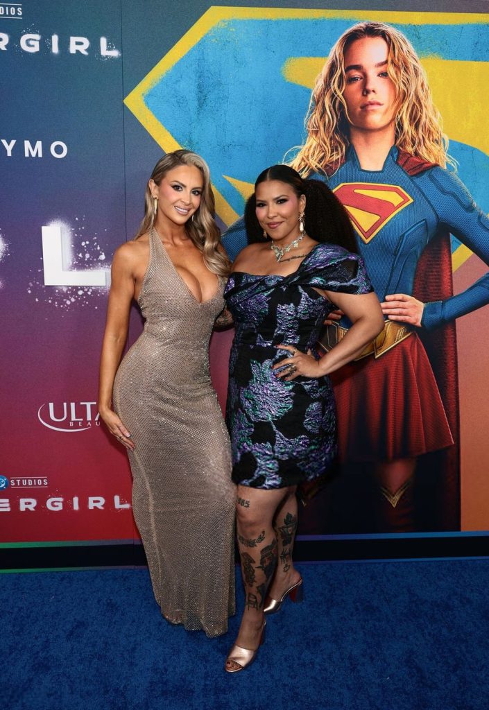 Two women pose together on a blue carpet at a “Supergirl” event. One wears a sparkly beige gown, the other a floral dress with tattoos visible. Behind them is a large “Supergirl” poster featuring the superhero.