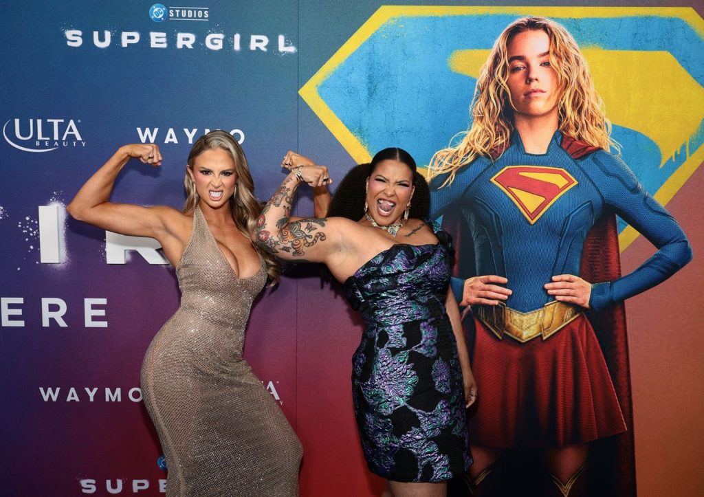 Two women pose, flexing their biceps and smiling, in front of a Supergirl poster featuring a woman in a Supergirl costume with her arms crossed. The event backdrop has logos and superhero-themed colors.