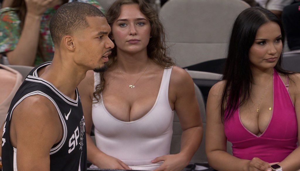 A basketball player in a black Spurs jersey stands on the court. Behind him, two women with long hair sit in the audience, one in a white top and the other in a pink top, both looking ahead.