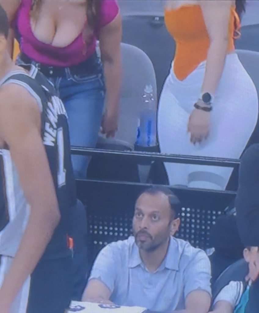 A man seated courtside at a basketball game looks upward with a surprised expression as two women stand in front of him, partially blocking his view.