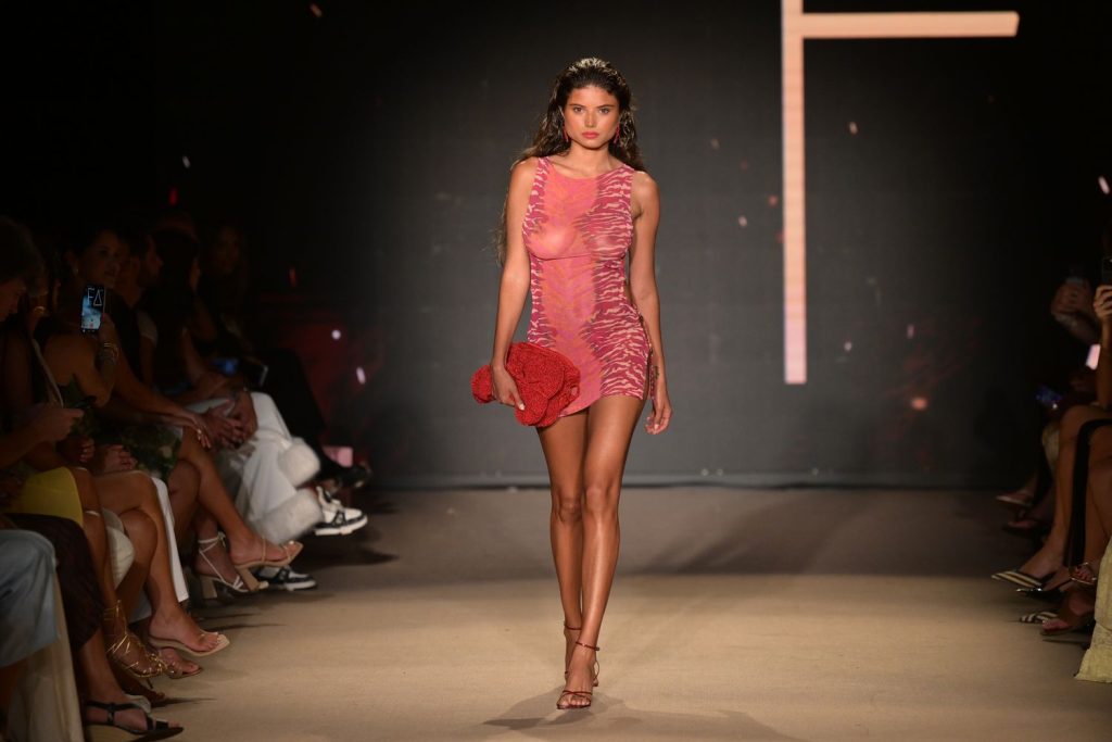 A model walks down a runway wearing a short, form-fitting, sleeveless pink dress with cutout details, holding a matching clutch. The audience is seated on both sides, and the background is dark with minimal lighting.