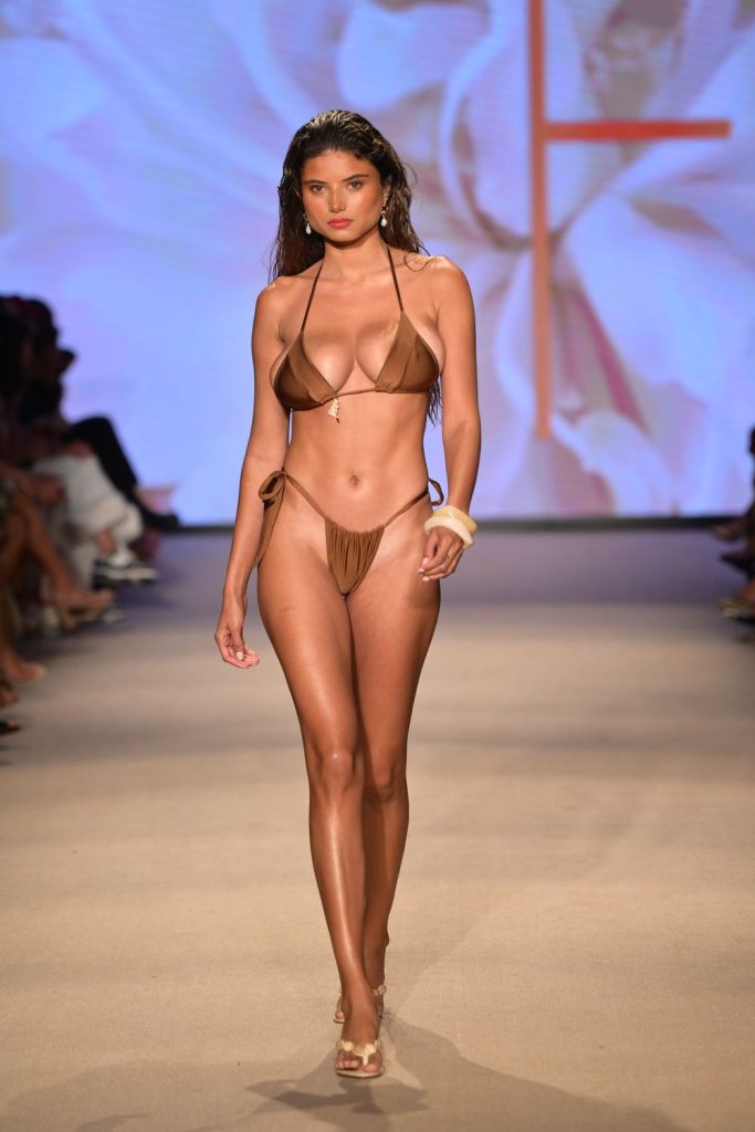 A model walks down a runway wearing a brown bikini with tie sides. The background features abstract floral graphics, and audience members sit on either side of the runway.