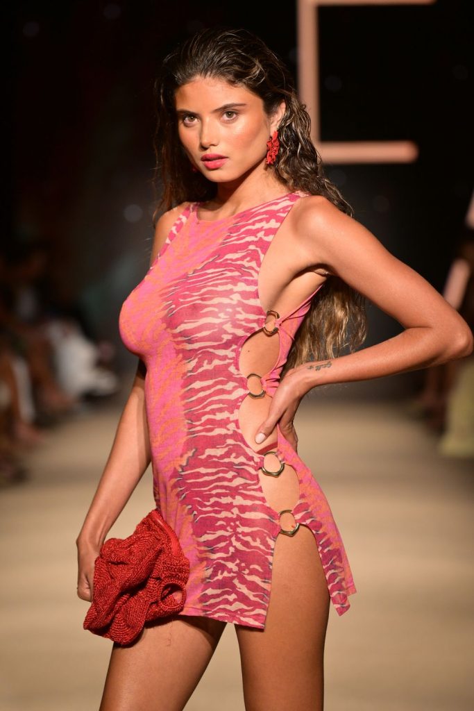 Close up of model with wavy dark hair stands at a side angle on the runway wearing a sleeveless, fitted pink and red patterned dress and holding a clutch.