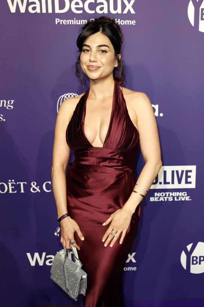 A woman in a deep red sleeveless gown poses on a purple carpet, holding a silver handbag. She has dark hair styled up, wears jewelry, and smiles at the camera. The background features event logos and white text.