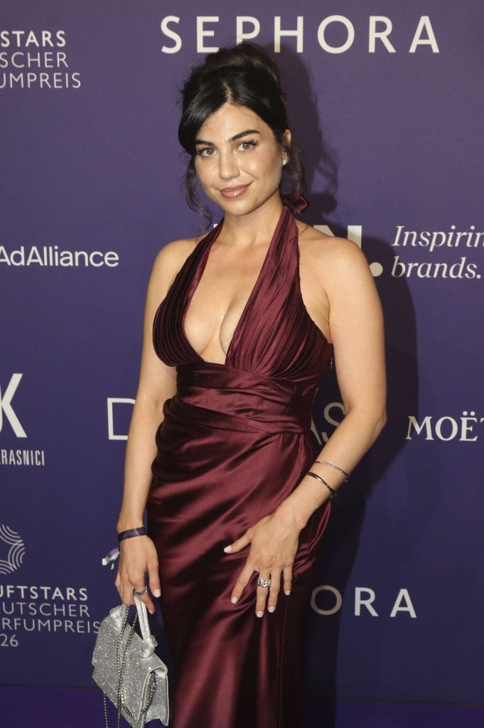 A woman with long dark hair in an elegant updo wears a deep burgundy satin evening gown with a plunging neckline, holding a silver handbag, posing on a purple carpet with a backdrop featuring various brand logos.