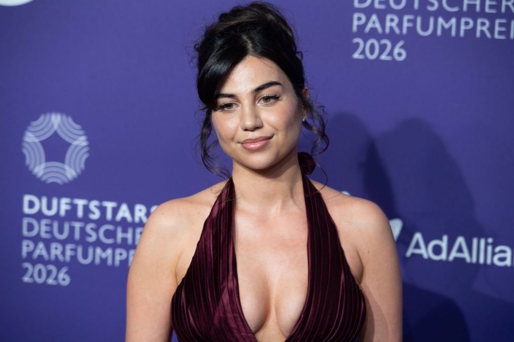 A woman with dark hair in an updo wears a deep burgundy, sleeveless dress with a plunging neckline, standing in front of a purple backdrop with white text and logos for a perfume event.