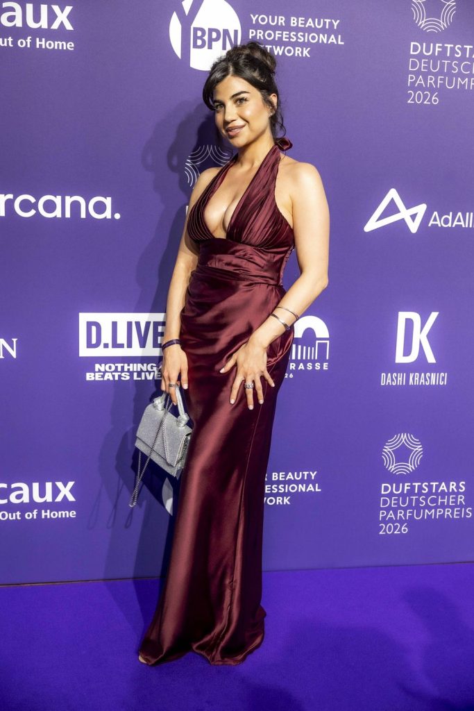A woman with dark hair in a low bun wears a deep burgundy halter gown, holding a silver bag, standing on a purple carpet with a purple backdrop featuring various logos and text.