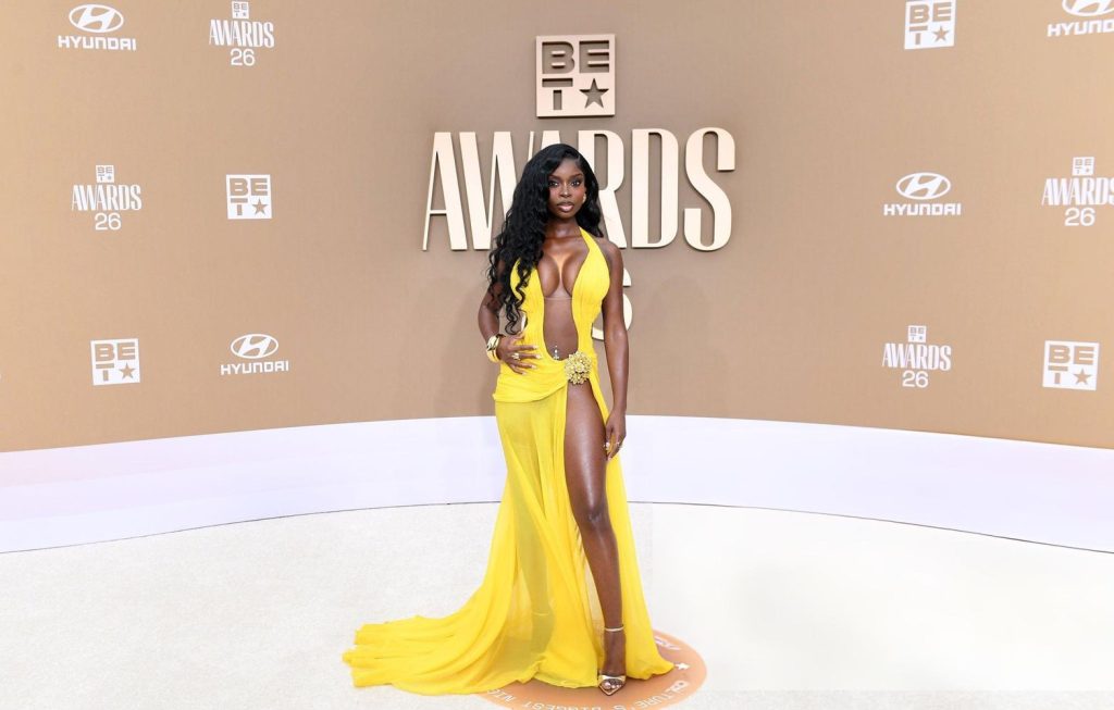 A woman in a bright yellow, flowing gown poses on the BET Awards red carpet. The dress features a high slit and deep neckline. The background displays BET Awards and Hyundai logos.