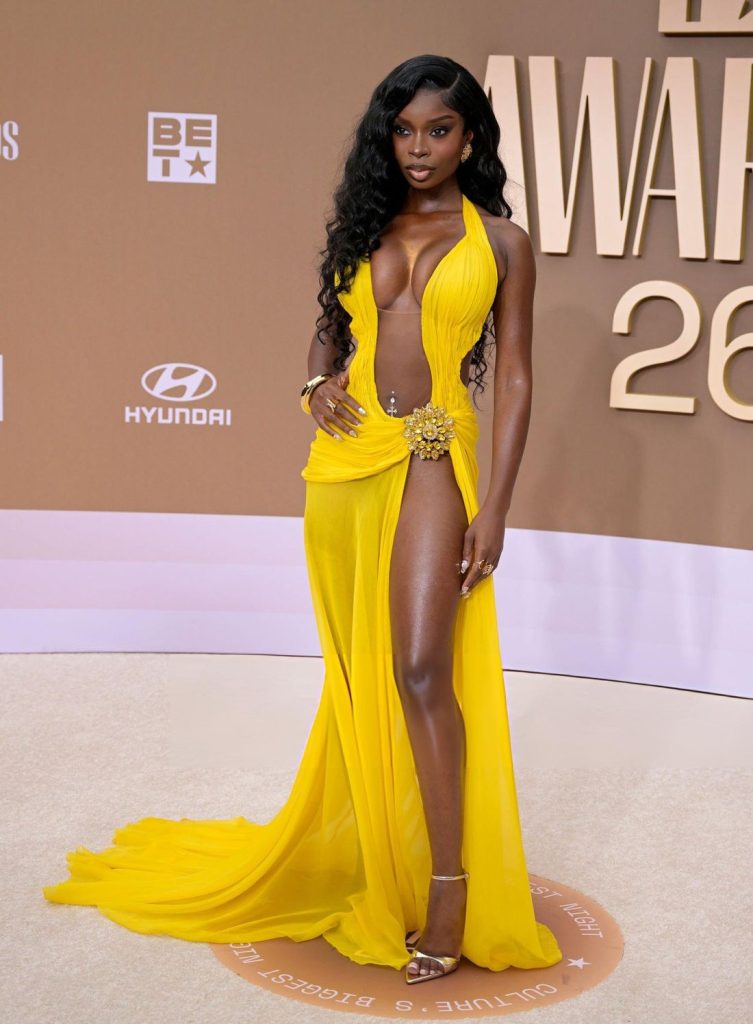 A woman poses on a beige carpet wearing a bright yellow, floor-length gown with a deep neckline, high slit, gold embellishment at the waist, and gold high heels. The background features BET Awards signage.