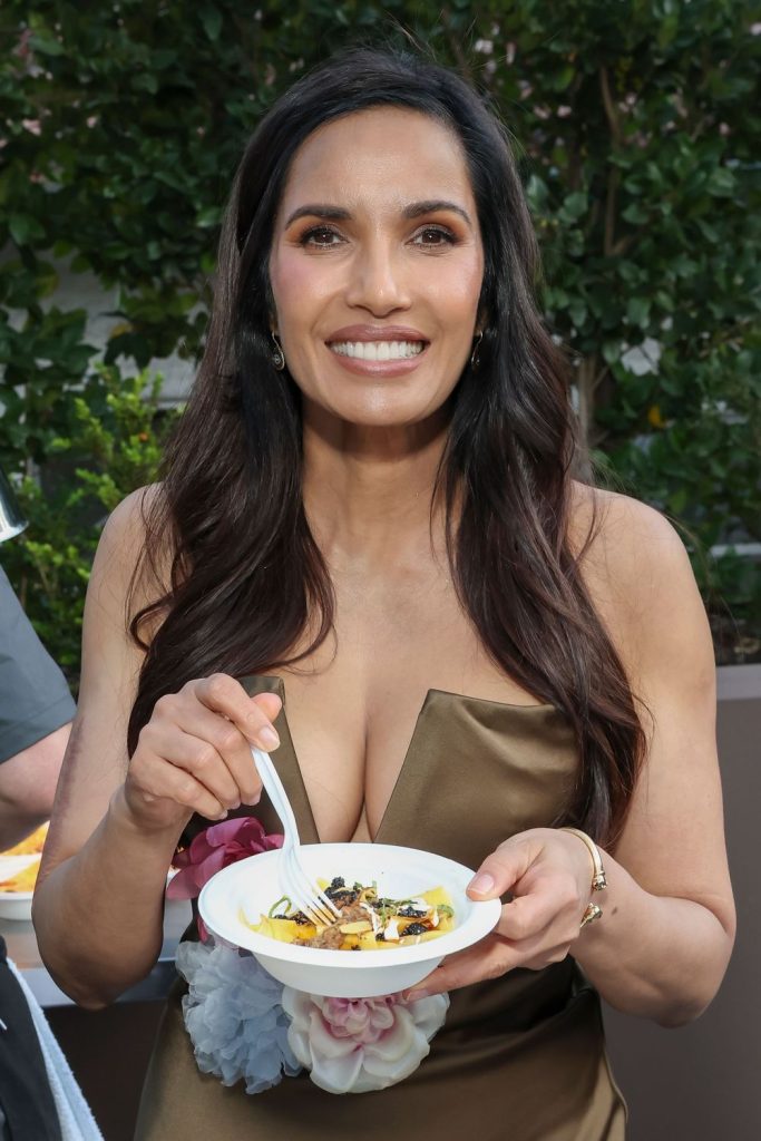 A woman with long dark hair in a bronze dress smiles while holding a white bowl of food and a fork, standing outdoors with greenery in the background.