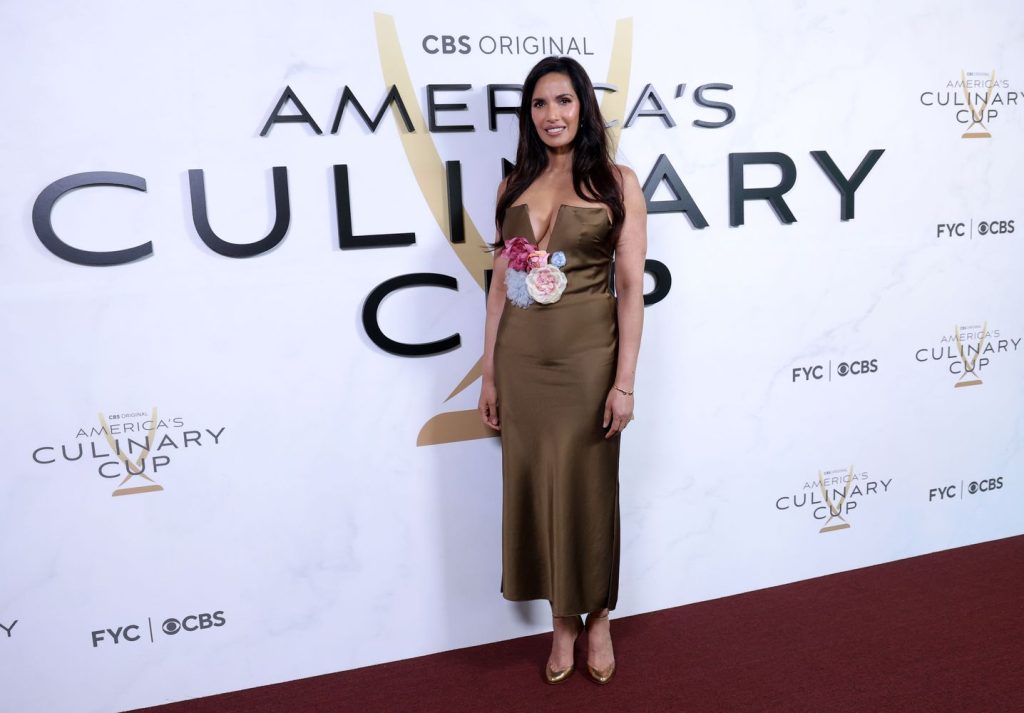A woman in a gold satin dress with flower details stands on a red carpet in front of a backdrop that reads Americas Culinary Cup and features CBS branding.