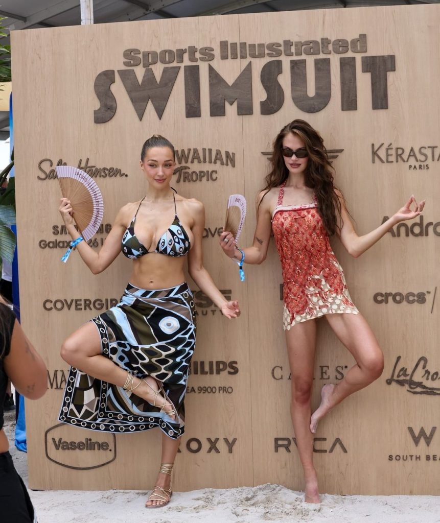Two women in colorful swimwear pose playfully on one leg, holding fans, in front of a large wooden board with Sports Illustrated Swimsuit and various brand logos at a beach event.