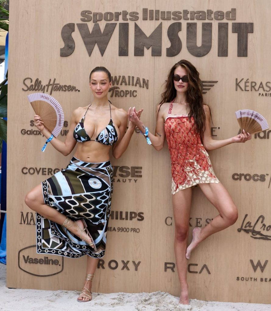Two women in colorful swimwear pose playfully on one leg, holding fans, in front of a large wooden board with Sports Illustrated Swimsuit and various brand logos at a beach event #3.