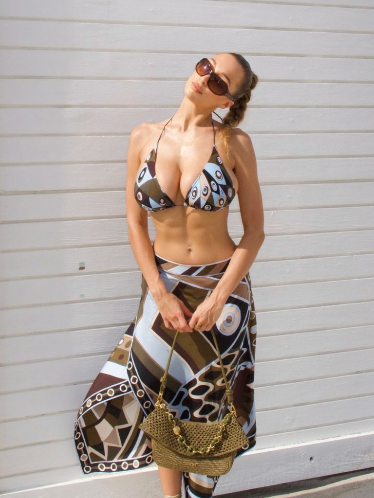 A woman wearing a patterned bikini and sarong stands in front of a white wooden wall on a sunny day. She has braided hair, sunglasses, strappy sandals, and holds a gold handbag.
