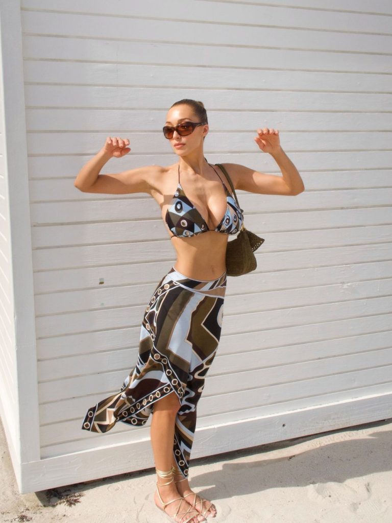 A woman wearing sunglasses, a patterned bikini top, and a matching skirt poses on a sandy beach in front of a white wooden wall, with her arms raised and a small bag on her shoulder.