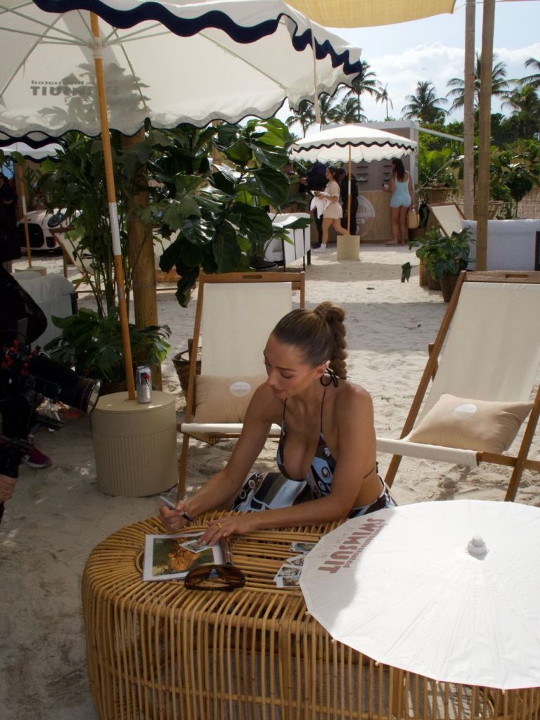 A woman in a swimsuit sits at a wicker table on a sandy beach, signing a magazine. She is surrounded by lounge chairs, plants, and umbrellas, with palm trees and other people in the background.