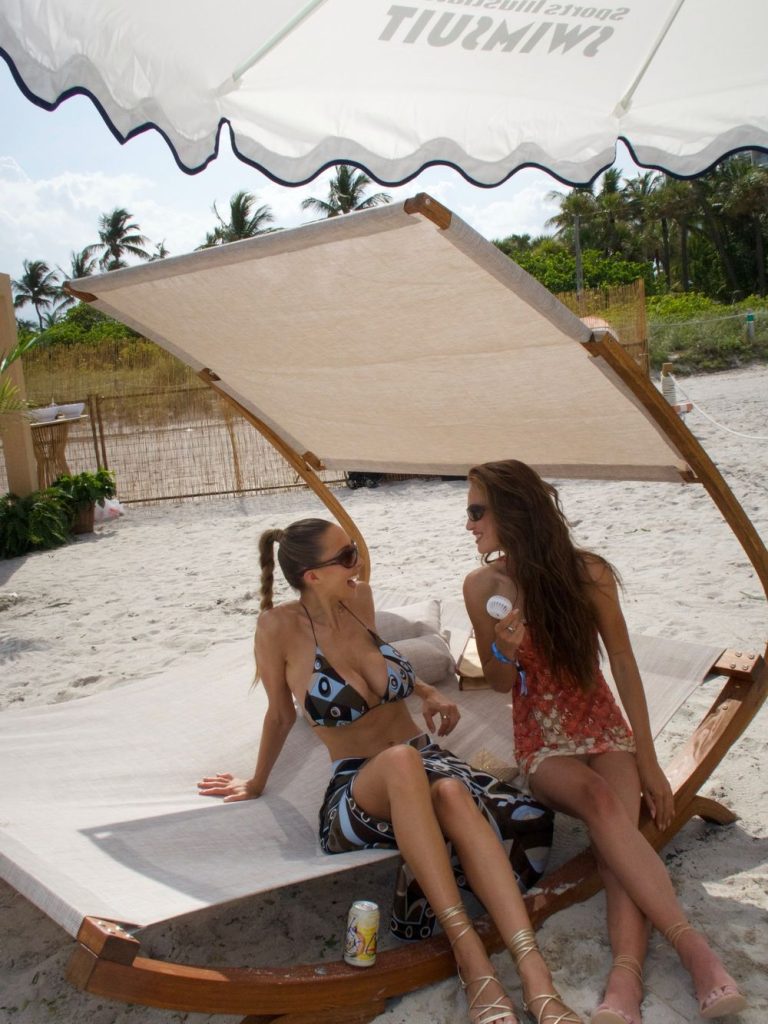 Two women in summer outfits sit and talk on a wooden beach lounge chair under a sunshade; one wears a patterned bikini and skirt, the other a red floral cover-up. There are palm trees and sand in the background.