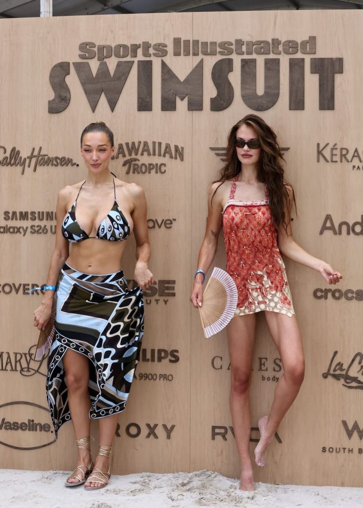 Two women pose in swimwear in front of a wooden wall with the Sports Illustrated Swimsuit logo and sponsor names. One wears a geometric bikini and skirt, the other a printed cover-up and holds a fan. They stand barefoot on sand.